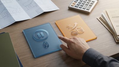 Hand points to blue 'pause/zero' financial deferral card next to orange 'percentage/coins' card on a desk with blueprints.