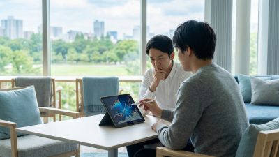 Two individuals analyze financial graphs on a tablet in a bright modern office with a city view. One points, the other contemplates.