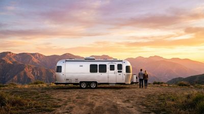 Sleek silver motorhome on mountain overlook. Two people watch a vibrant orange, pink, purple sunset over majestic peaks.
