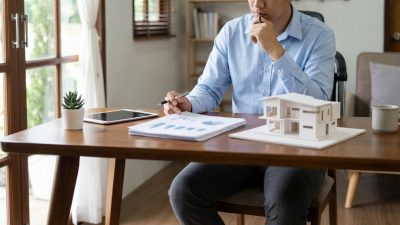 Indivíduo em camisa azul analisa documentos e maquete de casa em mesa de madeira, planejando aquisição de imóvel.
