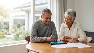 Diverse senior couple smiles, reviewing documents and a tablet at a bright home table, planning their secure future.