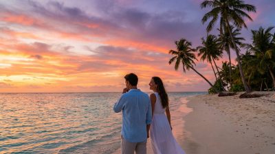 Couple on a pristine tropical beach at sunset. Sky ablaze with orange, pink, purple hues reflects on the calm ocean. Palm trees sway. One pensive, one smiling.
